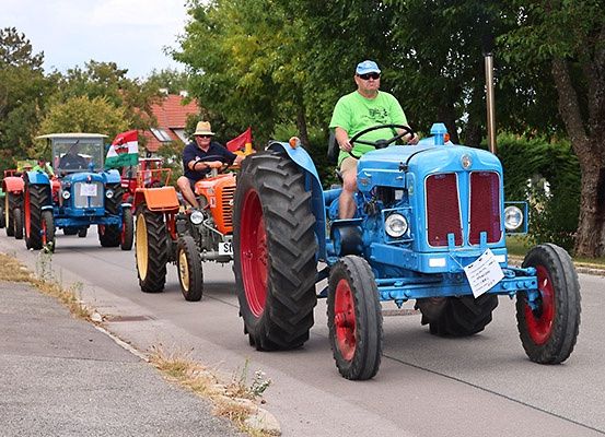 Eine Parade von blauen und orangefarbenen Traktoren fährt eine Landstraße hinunter, mit Bäumen und einem Haus im Hintergrund.