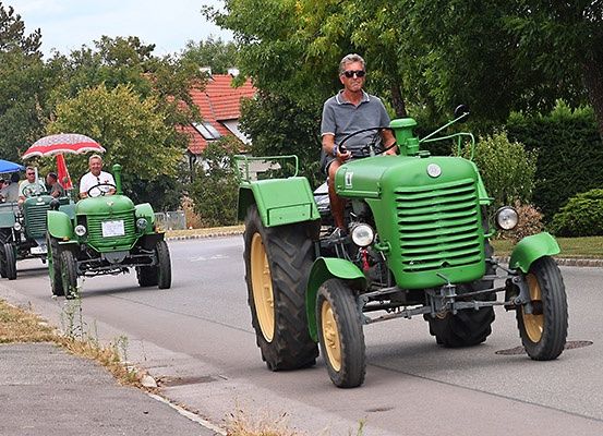 Ein Mann fährt einen grünen Traktor die Straße entlang, mit einem weiteren Traktor dahinter. Über einem der Traktoren befindet sich ein Regenschirm. Bäume und ein Haus sind im Hintergrund.