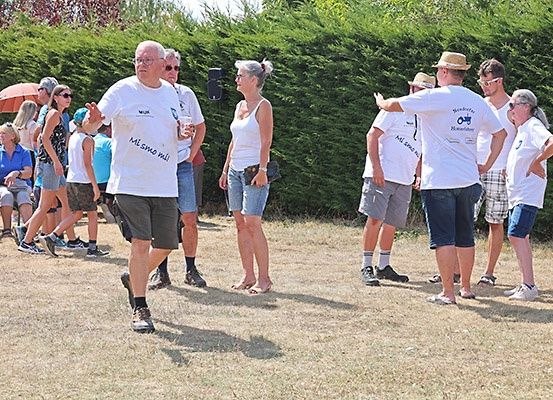 Eine Gruppe von Menschen versammelt sich im Freien. Einige tragen Hüte, Brillen und Turnschuhe. Einer hält ein Getränk. Sie stehen im Kreis, mit einer Hecke und Grünpflanzen hinter ihnen.