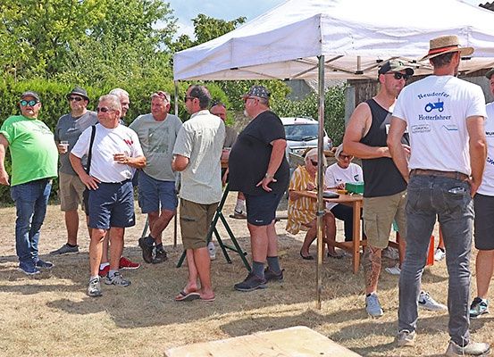Eine Gruppe von Männern versammelt sich unter einem weißen Zelt in einer Außenumgebung, einige tragen Hüte und Sonnenbrillen. Ein Tisch mit Gegenständen steht unter dem Zelt, und ein Auto ist in der Nähe geparkt.