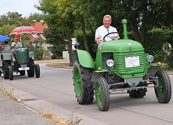 Zwei Oldtimer-Traktoren fahren eine Straße entlang, einer mit einem Mann unter einem Regenschirm und der andere mit einem lächelnden Fahrer. Bäume und Häuser sind im Hintergrund.