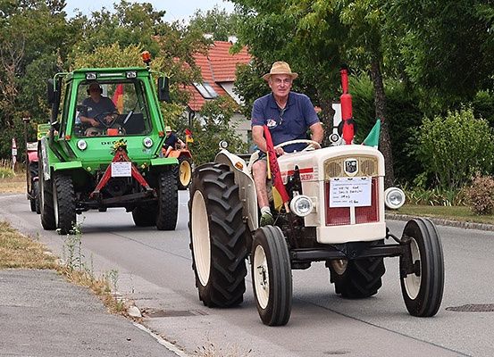Eine Gruppe von Landwirten fährt mit Traktoren in einer Parade. Ein Mann fährt einen weißen Traktor, während ein anderer Mann einen grünen Traktor fährt. Die Traktoren sind mit Flaggen geschmückt und ein Haus ist im Hintergrund zu sehen.