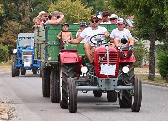 Eine Gruppe von Menschen, einschließlich Kindern, fährt auf einem roten Traktor, während ein blauer Traktor auf einer Straße mit Bäumen auf beiden Seiten folgt.