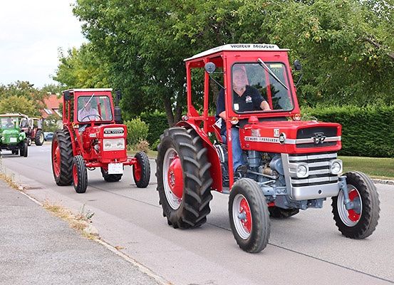 Mehrere rote Traktoren fahren auf der Straße. Eine Person sitzt auf dem Vordersitz des Traktors. Hinter dem Traktor befinden sich grüne Fahrzeuge.