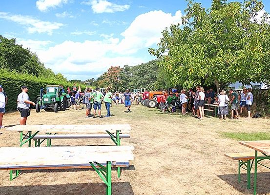 Ein sonniger Tag, mehrere Menschen sind um grüne Traktoren auf einem Feld versammelt, mit Picknicktischen und Bäumen im Hintergrund.