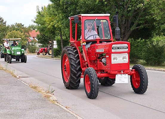 Ein roter Traktor fährt mit einem grünen Traktor dahinter auf der Straße, umgeben von Bäumen und einem Haus im Hintergrund.