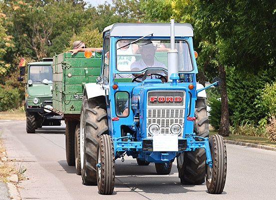 Ein blauer Ford-Traktor fährt auf der Straße und zieht einen grünen Anhänger hinter sich her. Eine Person sitzt auf dem Anhänger, während eine andere Person den Traktor fährt. Bäume stehen auf beiden Seiten der Straße.