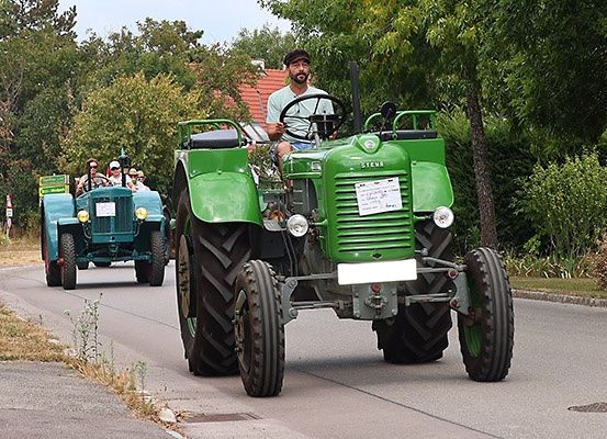 Zwei grüne Oldtimer-Traktoren fahren auf einer ländlichen Straße, wobei einer Passagiere transportiert und der andere Traktor hinterher fährt.