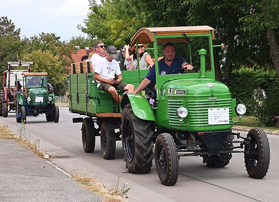 Eine Gruppe von Menschen fährt an einem sonnigen Tag auf einem grünen Oldtimer-Traktor, mit einem weiteren Traktor hinter ihnen auf einer ländlichen Straße.