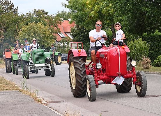 Eine Gruppe von Menschen fährt mit roten und grünen Traktoren eine Straße hinunter. Ein Traktor hat ein Kind darauf sitzen.