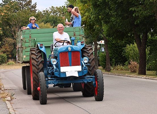 Ein älterer Mann fährt einen blauen Traktor mit einem Anhänger voller Menschen, die die Fahrt genießen, auf einer Straße.