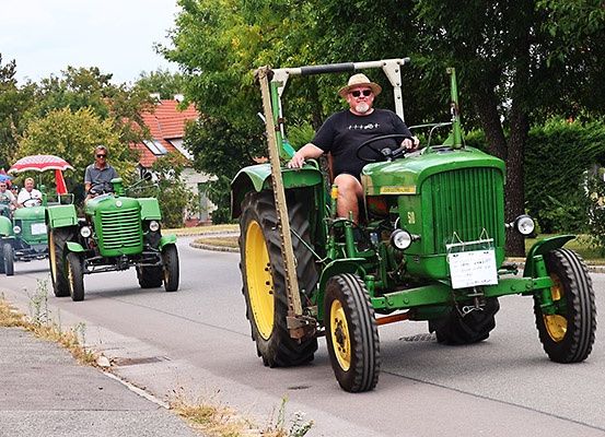 Eine Gruppe von Personen fährt mit grünen Traktoren auf einer Straße, mit Bäumen und Häusern im Hintergrund.