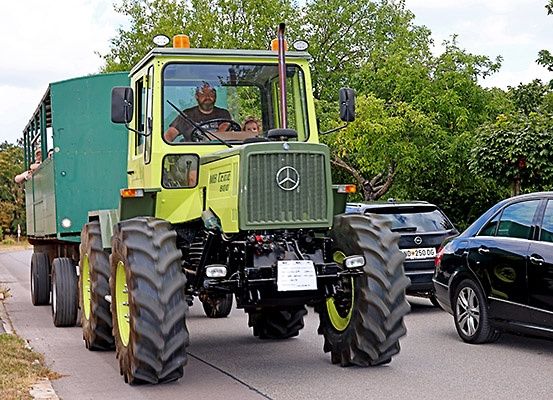 Ein gelber Traktor mit einem grünen Anhänger fährt eine Straße hinunter, hinter ihm fahren Autos.