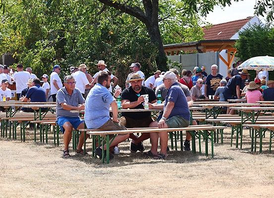 Eine Gruppe von Menschen sitzt auf Bänken in einem Außenbereich. Sie tragen alle Hüte und Sonnenbrillen. Einige von ihnen halten Flaschen und Dosen. Es stehen viele Menschen hinter ihnen.