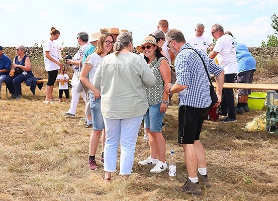 Eine Gruppe von Menschen, einige stehend und andere sitzend, versammelt sich auf einem Feld, lächelt und unterhält sich. Sie tragen Freizeitkleidung und Sonnenbrillen. Es gibt einige Flaschen und einen grünen Eimer auf einem Tisch.