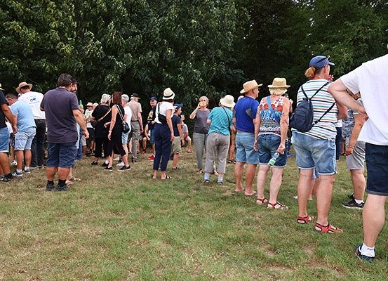 Eine Gruppe von Menschen steht in einer Reihe auf einem Rasen, wahrscheinlich auf etwas wartend. Die meisten tragen Hüte und Sandalen. Bäume stehen hinter ihnen.