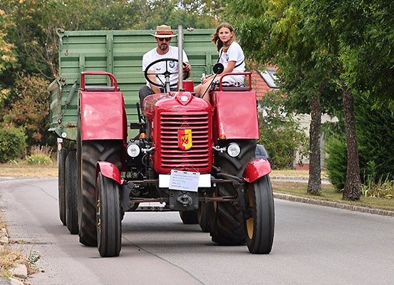Ein Mann und eine Frau fahren einen roten Traktor mit einem grünen Anhänger auf einer von Bäumen gesäumten Straße.