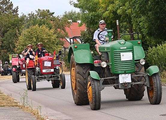 Ein Konvoi von Oldtimer-Traktoren fährt eine Landstraße hinunter, angeführt von einem Mann, der einen grünen Traktor fährt, gefolgt von einem roten Traktor mit der Nummer 1958.