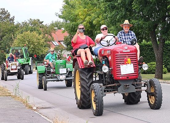 Mehrere Menschen fahren auf Traktoren in einer Parade, eine Frau macht ein Foto. Sie sind auf einer Straße, gesäumt von Bäumen und Häusern.
