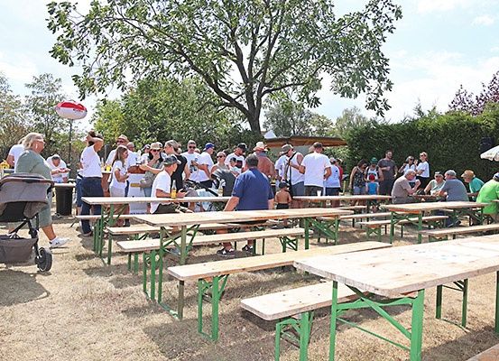 Eine Gruppe von Menschen sitzt an Picknicktischen in einem Außenbereich, einige stehen. Es gibt Bäume, eine Hecke und einen Kinderwagen in der Szene.