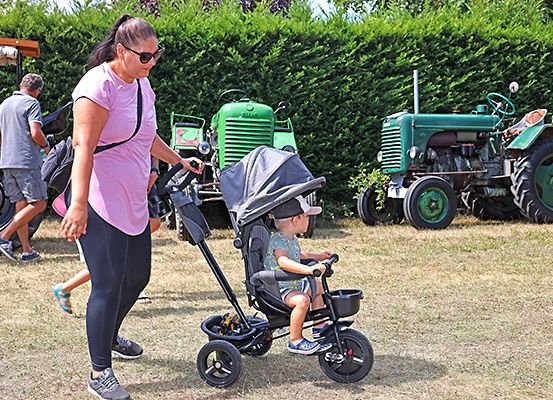 Eine Frau schiebt einen Kinderwagen mit einem Kind, das einen Dreirad-Kinderwagen fährt, vor einer Hecke und zwei Traktoren.