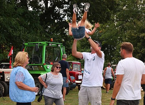 Eine Gruppe von Menschen steht draußen. Ein Mann hält ein kleines Mädchen in der Luft. Hinter ihnen sind Traktoren geparkt. Eine Frau auf der linken Seite hält ein Glas und eine andere Frau trägt eine Brille. Die Sonne scheint auf sie.