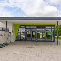 Das Äußere eines Kindergartens mit einer grünen Wand und Glastüren, einem Ziegelpflaster und einem bewölkten Himmel.