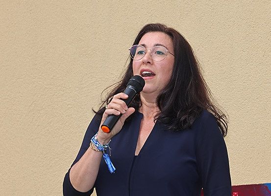 Eine Frau mit Brille und einem blauen Oberteil spricht in ein Mikrofon. Sie hält das Mikrofon mit der rechten Hand.