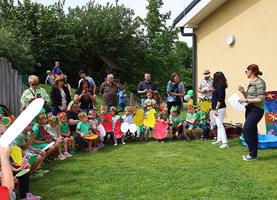 Eine Gruppe von Kindern und Erwachsenen versammelt sich im Freien für eine Veranstaltung, einige halten Requisiten, mit einem Gebäude und Bäumen im Hintergrund.