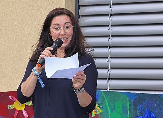 Eine Frau hält ein Mikrofon und Papier in der Hand, lächelt und spricht möglicherweise bei einer Veranstaltung. Sie trägt eine Brille und ein Armband. Hinter ihr befindet sich eine Wand mit Jalousien.