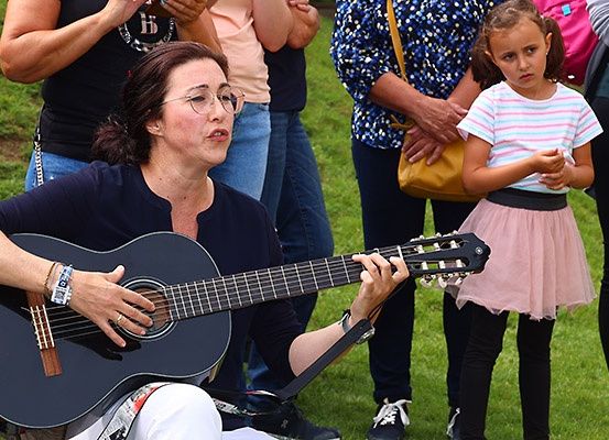 Eine Frau spielt Gitarre in einem Park mit einem kleinen Mädchen und mehreren Personen, die hinter ihr stehen.