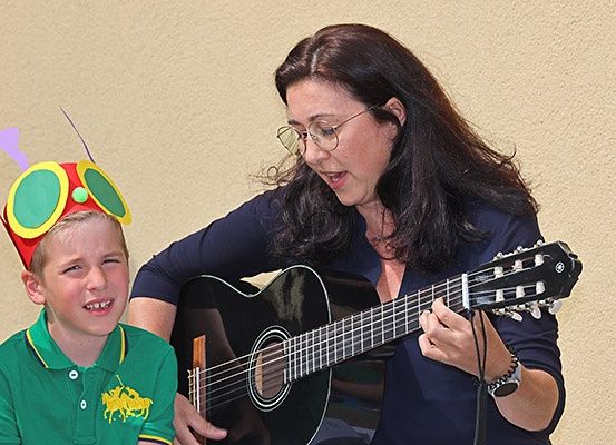 Eine Frau spielt Gitarre mit einem jungen Jungen, der eine Käfermütze trägt. Sie schauen beide in die Kamera.