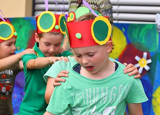Drei Kinder, die rote und gelbe Papierkronen tragen, führen ein Theaterstück auf. Ein Kind hält ein anderes in einem grünen T-Shirt mit den Worten 'Ich habe Hunger'.