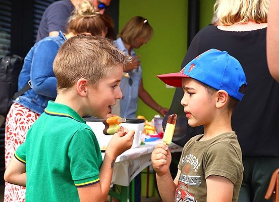 Zwei junge Jungen genießen Eis am Stiel bei einer Veranstaltung im Freien. Hinter ihnen gehen Leute umher, und ein Tisch mit verschiedenen Lebensmitteln ist zu sehen.