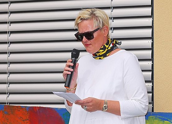 Eine Frau mit kurzen blonden Haaren spricht in ein Mikrofon, trägt ein weißes Oberteil und ein schwarz-gelbes Halstuch. In der anderen Hand hält sie ein Stück Papier, möglicherweise hält sie eine Rede. Hinter ihr befindet sich ein farbenfrohes Wandbild und ein weißer Fensterrollo.
