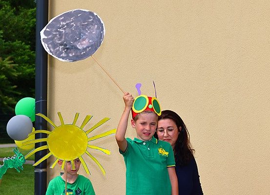 Zwei Jungen und eine Frau stehen im Freien. Ein Junge hält ein mondförmiges Papierbasteln. Sie sind von Luftballons und einer sonnenförmigen Dekoration umgeben. Bäume und ein Gebäude sind im Hintergrund.