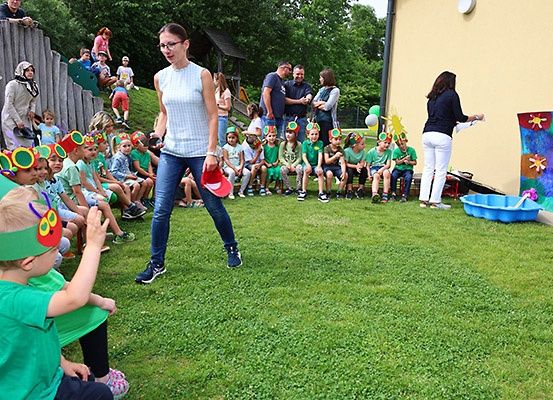 Kinder in grünen Kostümen sitzen auf dem Rasen, mit einer erwachsenen Frau, die einen roten Hut hält. Dahinter beobachtet eine Gruppe von Menschen, einige stehend und andere sitzend. Eine Frau malt eine bunte Wandmalerei an der Wand.