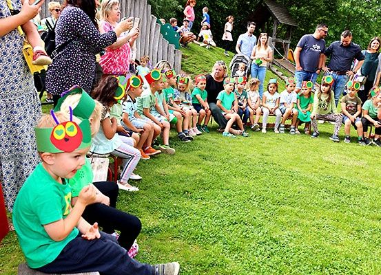 Kinder in grünen Hemden und Raupen-Kopfbedeckungen sitzen auf einer Rasenfläche mit Erwachsenen hinter ihnen. Einige Erwachsene halten Momente mit ihren Handys fest.