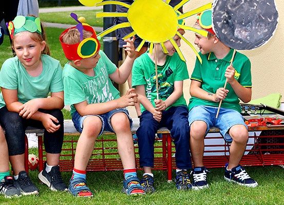 Vier Kinder sitzen auf einer Parkbank und tragen bunte Kopfbänder und Requisiten. Der Junge in der Mitte hält eine gelbe Sonne, während der Junge neben ihm einen Mond hält.