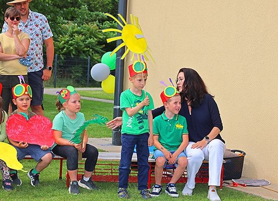 Eine Gruppe von Kindern und Erwachsenen versammelt sich im Freien, wobei mehrere Kinder Raupenmützen tragen und Papierausschnitte halten. Eine Frau interagiert mit einem der Kinder. Im Hintergrund befinden sich eine Wand, Luftballons und eine Sonnendekoration.