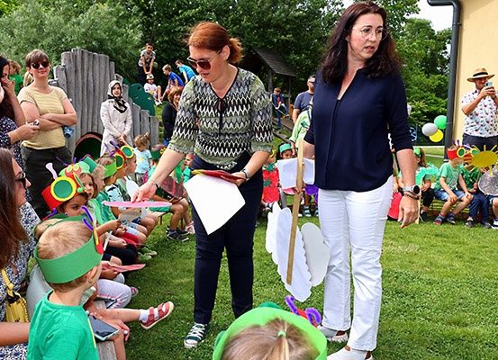 Zwei Frauen verteilen Papiere an Kinder, die als Raupen gekleidet sind, in einem Garten. Eine Frau hält einen Papierfalter.
