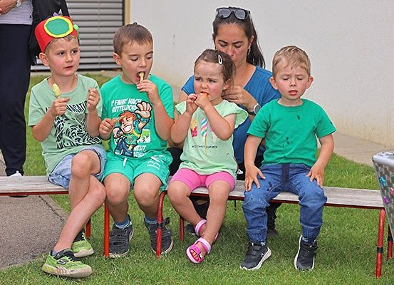 Vier Kinder und eine Frau sitzen auf einer Bank und essen Eispops. Die Kinder tragen grüne T-Shirts und rosa Röcke. Die Frau hält ein Kind auf dem Schoß.