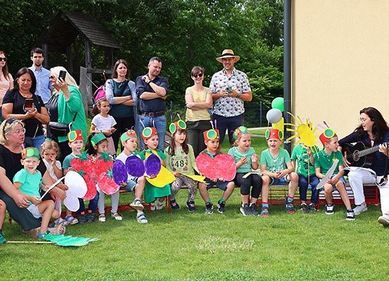 Eine Gruppe von Kindern und Erwachsenen versammelt sich im Freien, einige in Kostümen, während eine Frau auf einer Bank eine Gitarre spielt. Bäume und ein Gebäude sind im Hintergrund zu sehen.