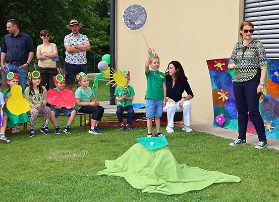 Eine Gruppe von Kindern und Erwachsenen nimmt an einer Outdoor-Veranstaltung teil. Kinder tragen Raupenhüte, während ein Kind einen Mondstab hält. Eine Erwachsene steht neben einer grünen Decke.