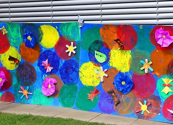 Ein farbenfrohes Wandbild zeigt eine Gartenszene mit bunten Kreisen, Blumen und Schmetterlingen. Der Hintergrund ist blau, und es ist an einer Wand mit Jalousien oben befestigt.