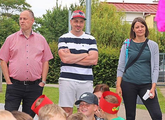 Eine Gruppe von Erwachsenen und Kindern versammelt sich draußen, möglicherweise für eine Veranstaltung. Ein Mann in einem rosa Hemd steht neben einem Mann in einem gestreiften Hemd, beide schauen in die Kamera. Eine Frau mit Rucksack hält ein Stück Papier. Kinder tragen Mützen.