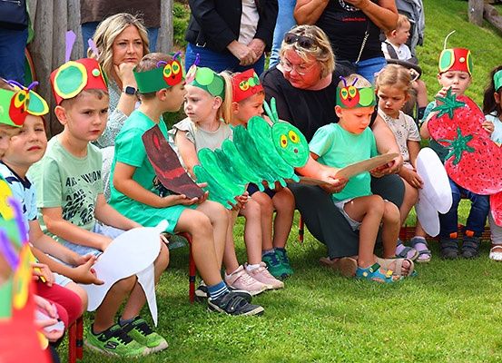 Kinder, die verschiedene Kostüme tragen, sitzen auf Stühlen im Freien. Sie tragen Mützen, die Raupen ähneln, und halten Requisiten. Eine Frau liest ihnen ein Buch vor.