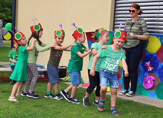 Eine Gruppe von Kindern in Kostümen tanzt im Freien, eine Frau beobachtet. Sie tragen bunte Hüte und grüne T-Shirts. Hinter ihnen befindet sich eine bemalte Wand mit Luftballons.