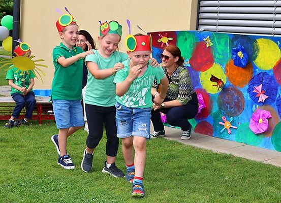 Vier Kinder mit Schmetterlingshüten gehen auf dem Rasen, während ein anderes auf einer Bank sitzt. Eine Frau hockt hinter ihnen neben einer bunten Wand.