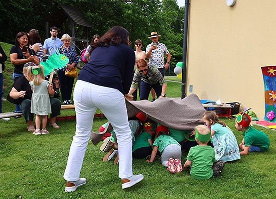 Mehrere Kinder spielen unter einer Decke auf dem Rasen. Einige Erwachsene stehen hinter ihnen und schauen zu. Das Gebäude hat Luftballons und ein Banner.